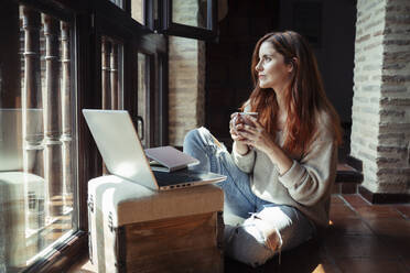 Junge Frau trinkt Kaffee, während sie zu Hause am Laptop arbeitet - JSMF01813