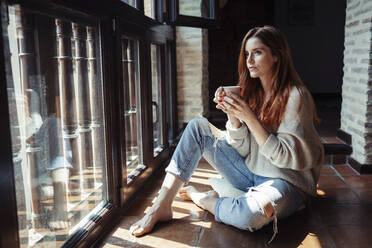 Young woman drinking coffee while sitting on floor at home - JSMF01814
