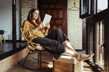 Smiling woman using digital tablet while sitting on chair at home - JSMF01815