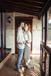 Smiling couple holding hands while standing by window at home - JSMF01837