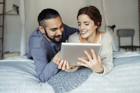 Lächelndes Paar, das ein digitales Tablet benutzt, während es zu Hause auf dem Bett liegt - JSMF01840
