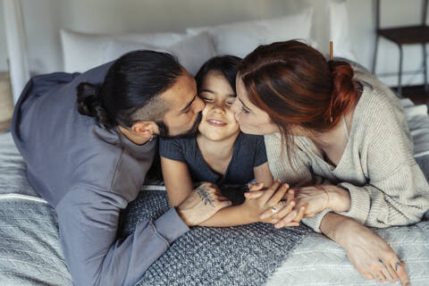 Mother and father kissing daughter while lying on bed at home stock photo