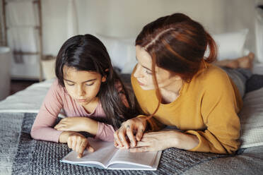 Daughter reading book while lying on bed by mother at home - JSMF01855