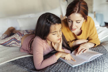 Girl and woman reading book while lying on bed at home - JSMF01857