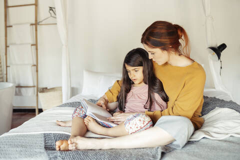 Mother and daughter reading book while sitting on bed at home stock photo