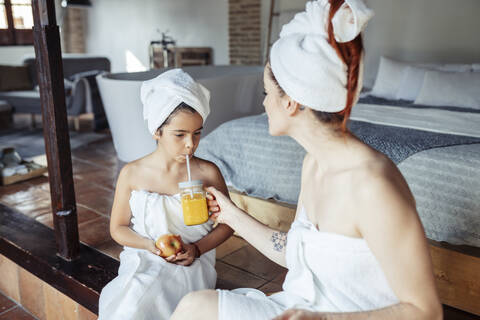 Mother in towel feeding juice to daughter while sitting at home stock photo