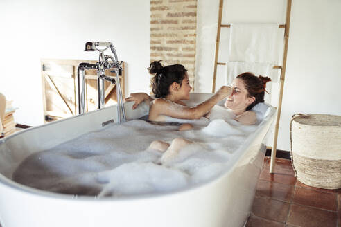 Mother and daughter playing while taking bath in bathtub at home - JSMF01877