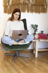Young woman sitting on chair while working on laptop at home - AFVF07327
