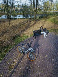 Aerial view of young woman sitting alone on bench in autumn park with pond in background - KNTF05727