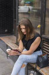 Junge Frau mit Smartphone und Einweg-Kaffeebecher auf einer Bank sitzend - BOYF01606