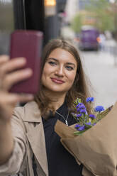 Lächelnde Frau, die ein Selfie mit einem Blumenstrauß macht, während sie auf einem Fußweg steht - BOYF01613