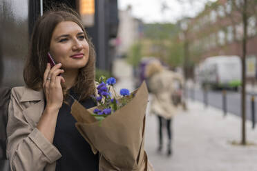 Junge Frau, die mit einem Smartphone telefoniert und wegschaut, während sie einen Blumenstrauß hält - BOYF01614