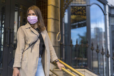 Young woman wearing protective face mask standing against door during covid-19 - BOYF01644