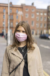 Young woman wearing protective face mask standing on street in city during covid-19 - BOYF01646