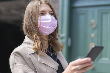 Young woman using smart phone while wearing protective face mask standing during covid-19 - BOYF01647