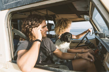 Young woman sitting by dog and man while driving car at beach - DAMF00557