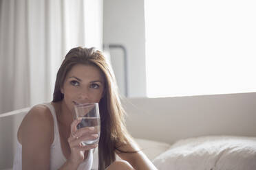 Young woman drinking water from glass in bedroom at home - AJOF00241