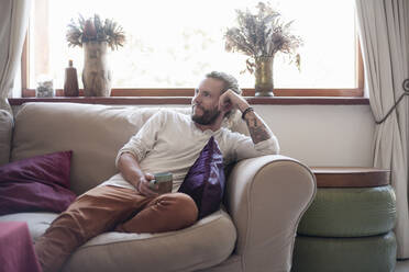 Male hipster having coffee while relaxing on sofa at home - AJOF00249