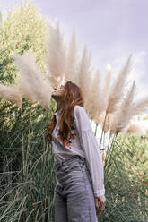 Young redhead woman standing by pampas grass in garden - AFVF07329