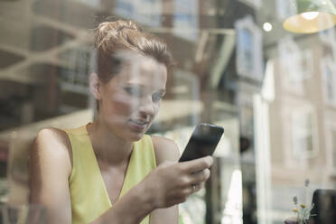 Lächelnde Geschäftsfrau, die ein Smartphone in einem Café benutzt, gesehen durch ein Glasfenster - AJOF00261