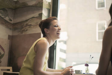 Young businesswoman laughing while discussing with female friend during meeting in coffee shop - AJOF00266