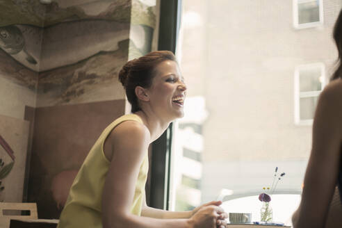 Junge Geschäftsfrau lacht, während sie mit einer Freundin während eines Treffens in einem Café diskutiert - AJOF00266
