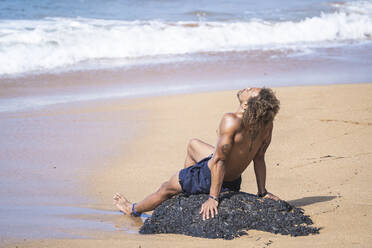 Young shirtless man relaxing on rock at beach - SNF00626