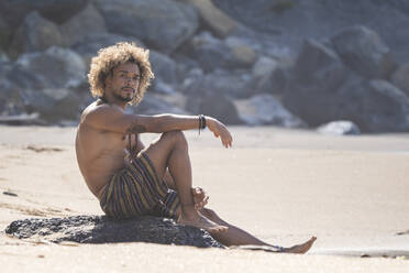 Hemdloser junger Mann sitzt auf einem Felsen am Strand im Sommerurlaub - SNF00627