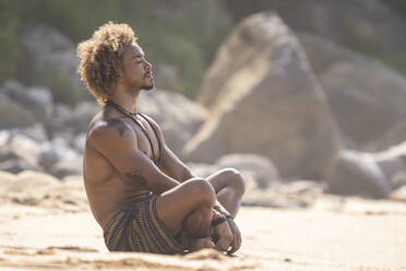 Hemdloser junger Mann meditiert am Strand an einem sonnigen Tag - SNF00629