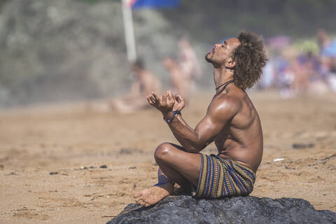 Shirtless young man gesturing while meditating on rock at beach during summer vacation stock photo