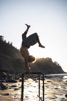 Hemdloser junger Mann übt Handstand am Barren am Strand - SNF00658