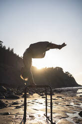 Hemdloser junger Mann übt Handstand am Barren am Strand bei Sonnenuntergang - SNF00659