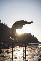 Shirtless young man practicing handstand on parallel bars at beach during sunset - SNF00659