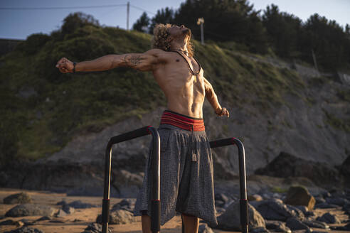 Shirtless muscular man standing with arms outstretched at beach - SNF00660