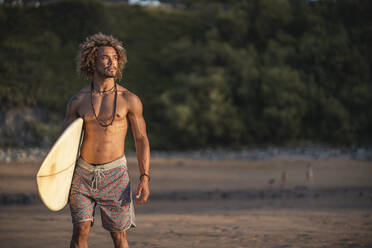 Hemdloser junger Mann mit Surfbrett, der bei einem Strandspaziergang bei Sonnenuntergang wegschaut - SNF00665