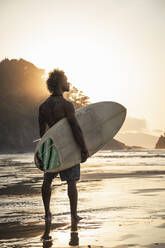Hemdloser junger Mann mit Surfbrett steht am Meer gegen den Himmel bei Sonnenuntergang - SNF00671