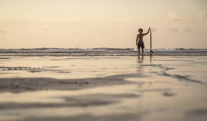 Shirtless man standing with surfboard at seashore against sky during sunset - SNF00674