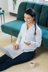 Junge Frau mit Laptop, Smartphone und Kaffeetrinken zu Hause sitzend - GIOF09158