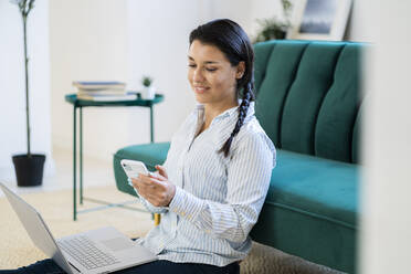 Junge Frau mit Laptop, die zu Hause sitzend eine SMS auf ihrem Smartphone schreibt - GIOF09159