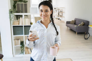 Smiling woman using smart phone while holding coffee cup standing at home - GIOF09161
