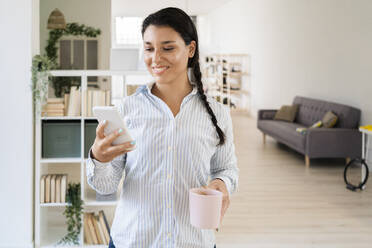 Smiling woman text messaging on smart phone while holding coffee cup standing at home - GIOF09163