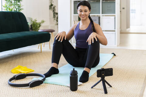 Smiling young woman video recording while sitting on exercise mat at home stock photo