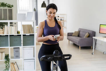 Smiling young woman using smart phone while sitting on exercise bike at home - GIOF09189