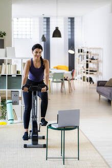 Weibliche Sportlerin trainiert auf einem Heimtrainer, während sie zu Hause einen Laptop benutzt - GIOF09192