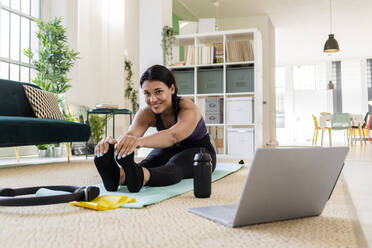 Smiling female athlete doing stretching exercise while sitting at home - GIOF09213