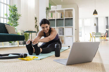 Smiling young woman looking away while doing stretching exercise sitting at home - GIOF09215