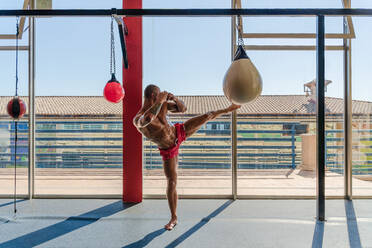 Seitenansicht eines muskulösen männlichen Kickboxers mit nacktem Oberkörper, der während eines intensiven Trainings in einem Fitnessstudio Kicks am Boxsack übt - ADSF16638