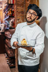 Seriöser männlicher Koch in Uniform steht mit leckeren Snacks auf einer Schiefertafel im Café und schaut in die Kamera - ADSF16643