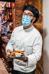 Seriöser männlicher Koch in Uniform und medizinischer Maske steht mit leckeren Snacks auf einer Schiefertafel in einem Café und schaut in die Kamera - ADSF16644