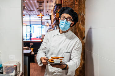 Seriöser männlicher Koch in Uniform und medizinischer Maske steht mit leckeren Snacks auf einer Schiefertafel in einem Café und schaut in die Kamera - ADSF16645